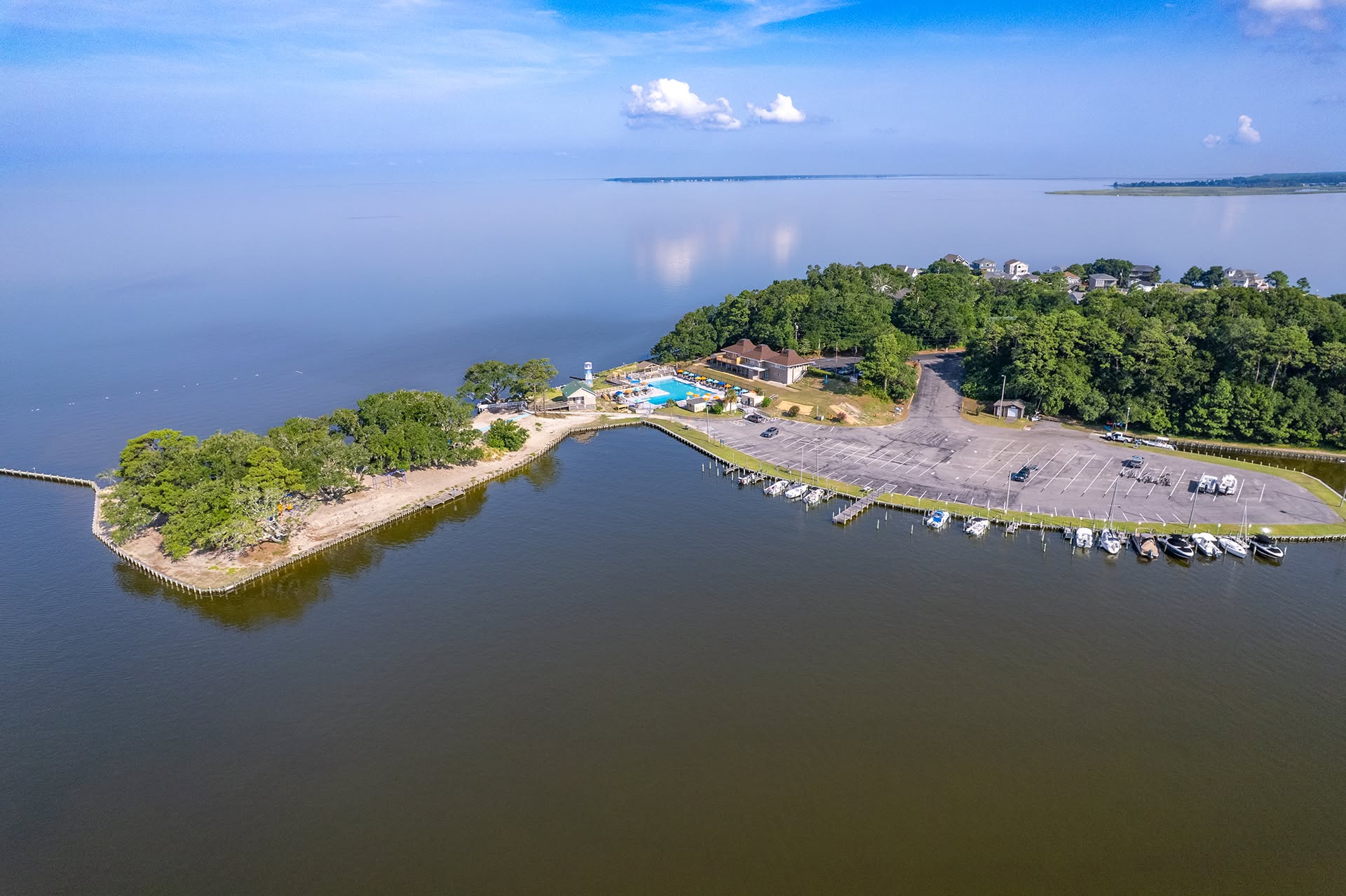 Aerial view of a peninsula with a parking lot, trees, and a small marina by a calm body of water.