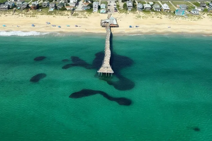 Aerial view of a pier extending into the ocean from a sandy beach with nearby houses.