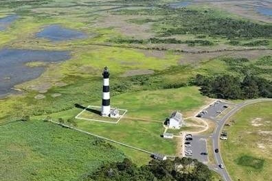 Aerial view of a striped lighthouse on coastal landscape with nearby roads and trees.