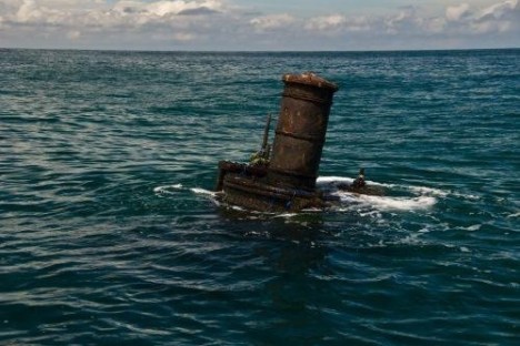 Rusty chimney partly submerged in ocean water with clouds in the sky.