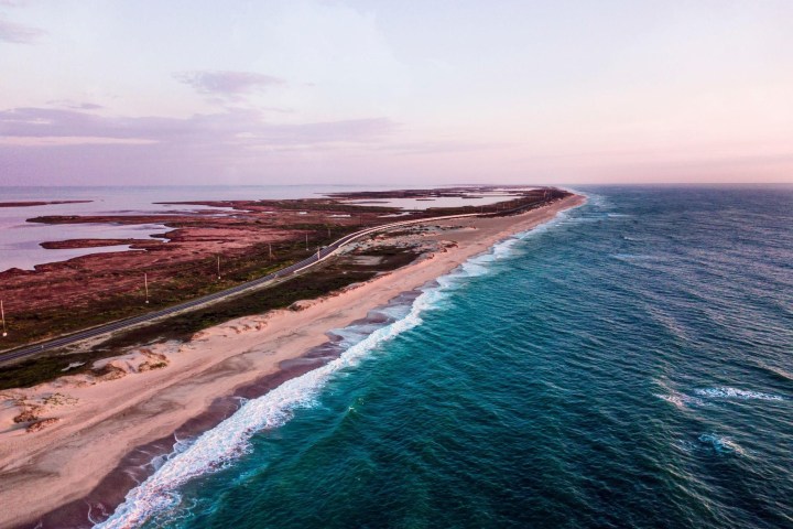 Aerial view of a coastal road between a sandy beach and the ocean at sunset.